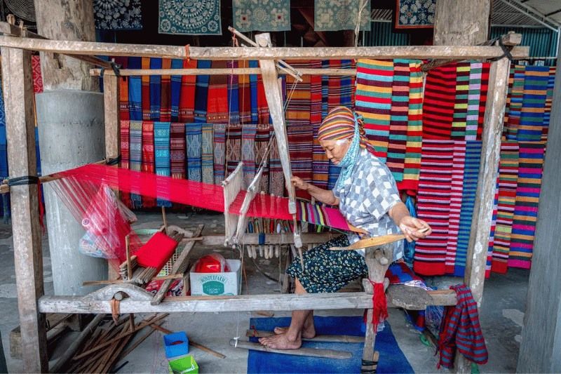 Ältere frau webt bunte stoffe an einem webstuhl auf einem lebhaften dorfmarkt und bewahrt traditionelle handwerkskunst (quelle quang nguyen vinh - pexels)